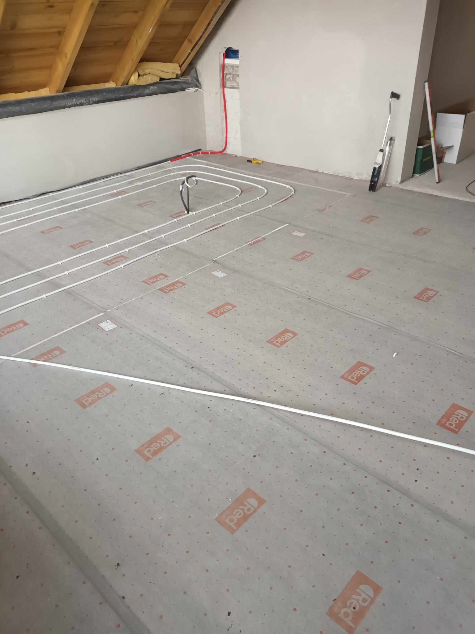 Białe rury ogrzewania podłogowego ułożone na izolacji z nadrukowanym logo 'Red' w surowym wnętrzu poddasza ze skośnym sufitem.