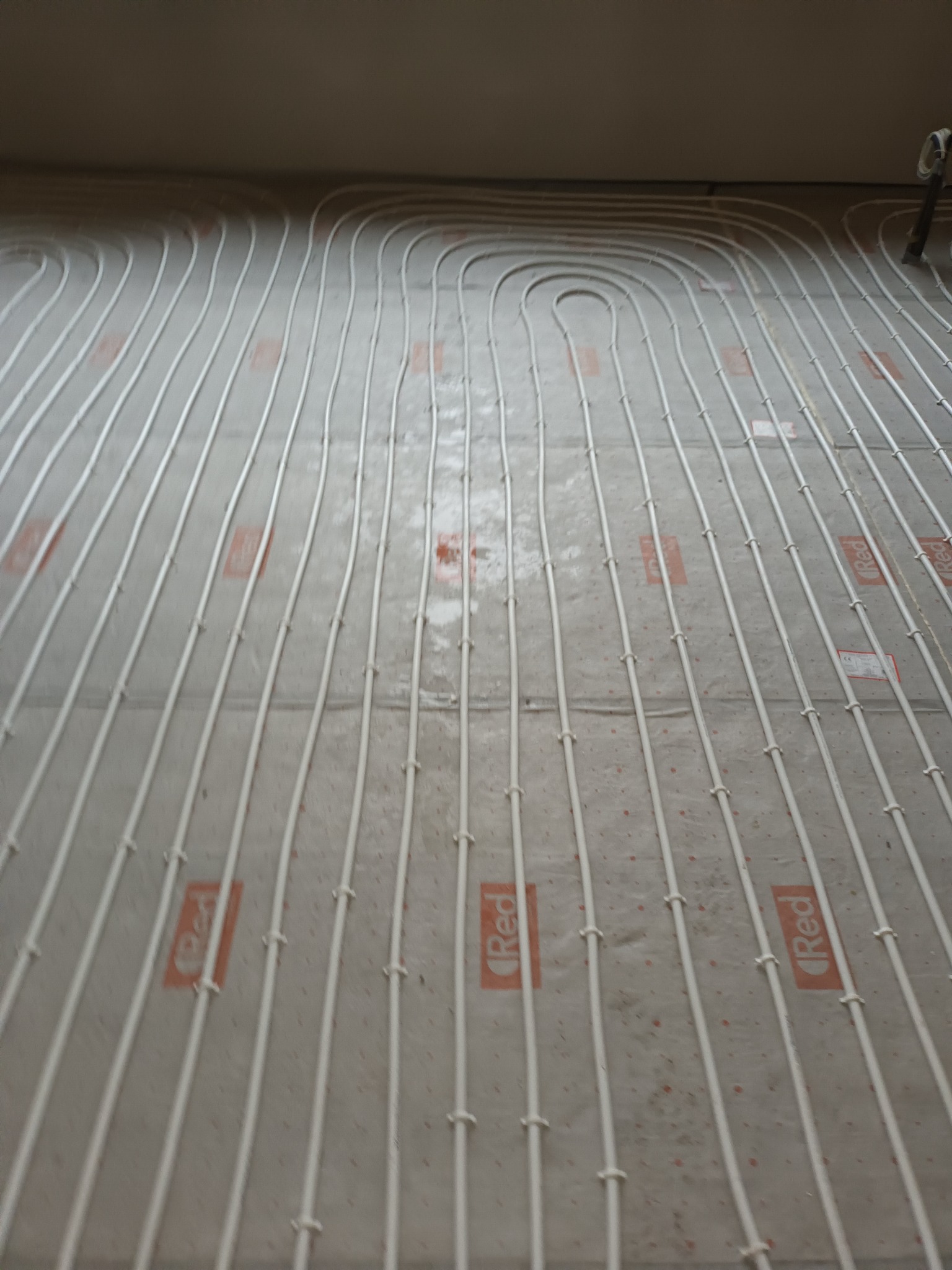 Białe rurki ogrzewania podłogowego ułożone na szarej macie z pomarańczowymi oznaczeniami 'Red', widok z góry w trakcie montażu.