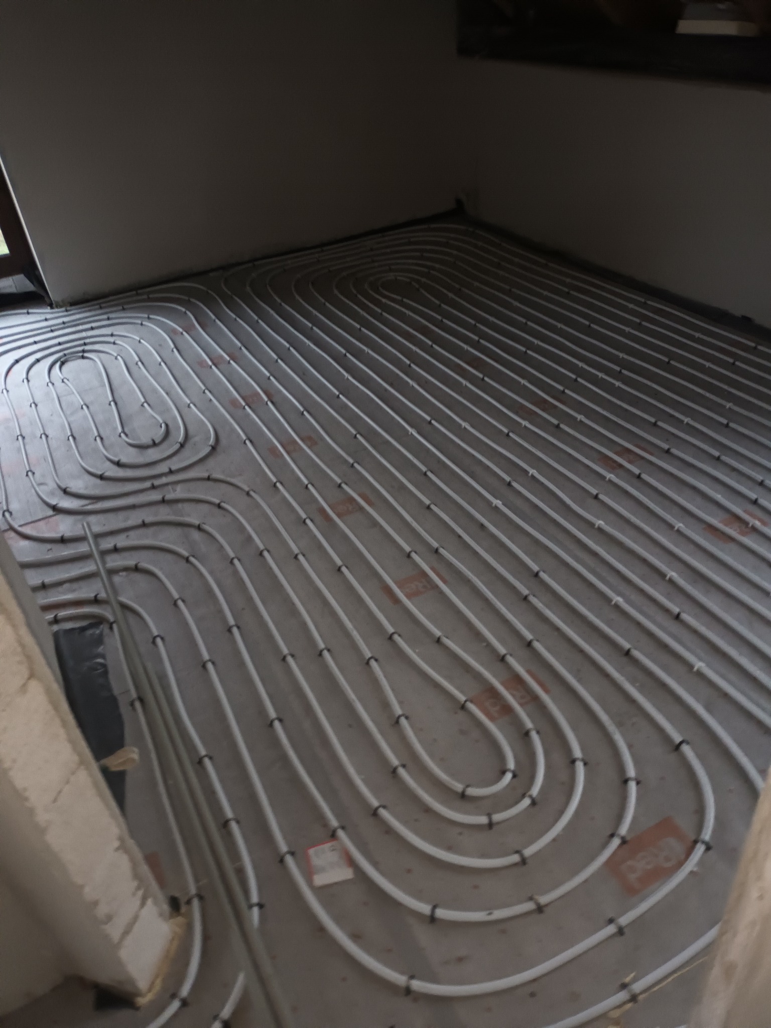 Białe rury układane spiralnie na szarym podłożu, tworzące system grzewczy podłogowy, widoczne czarne mocowania rur, surowy stan wykończenia, widok z góry.