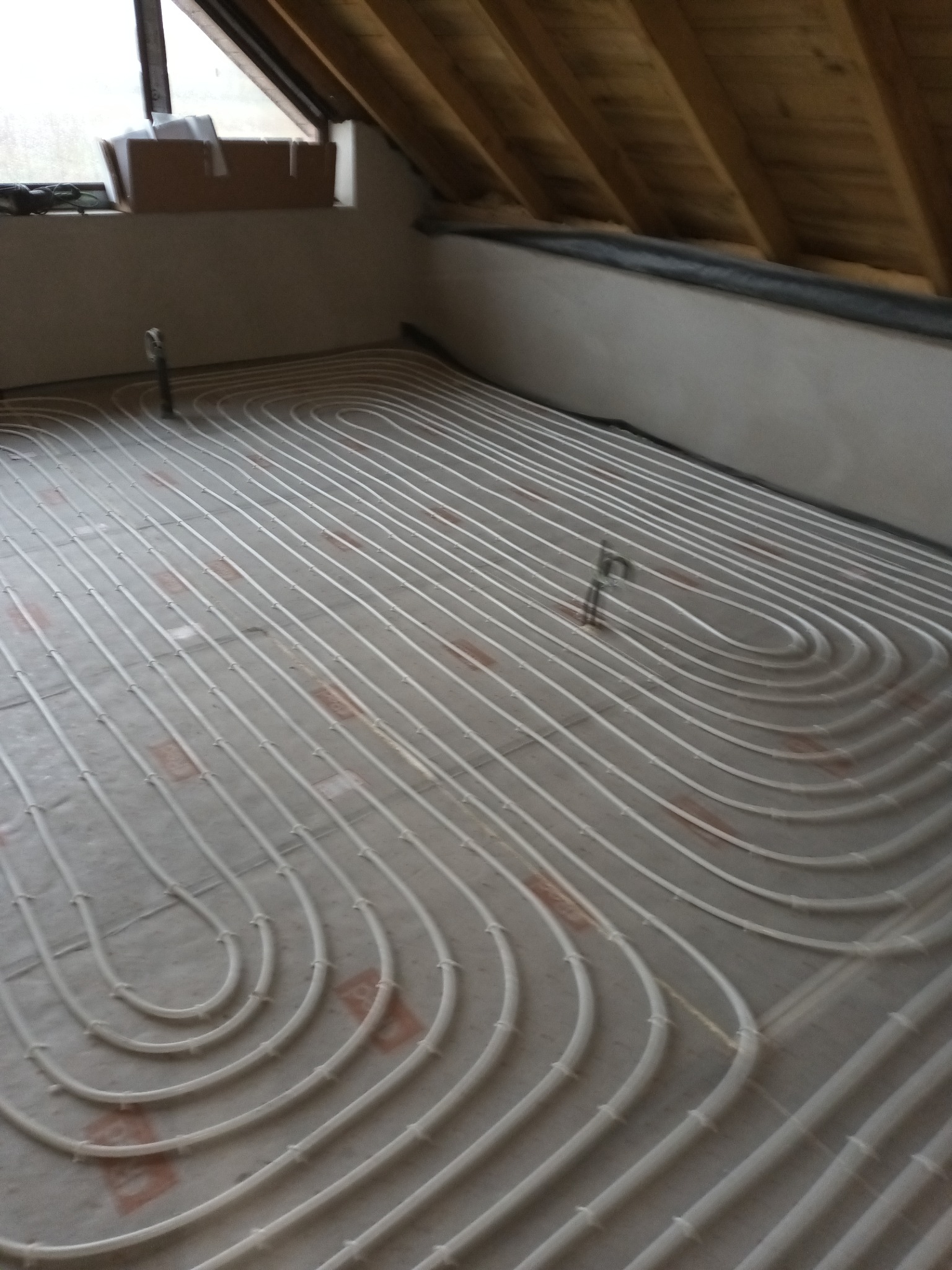 Białe rury ogrzewania podłogowego ułożone w pętle na szarym podłożu w pomieszczeniu na poddaszu, widoczne drewniane elementy konstrukcyjne dachu.