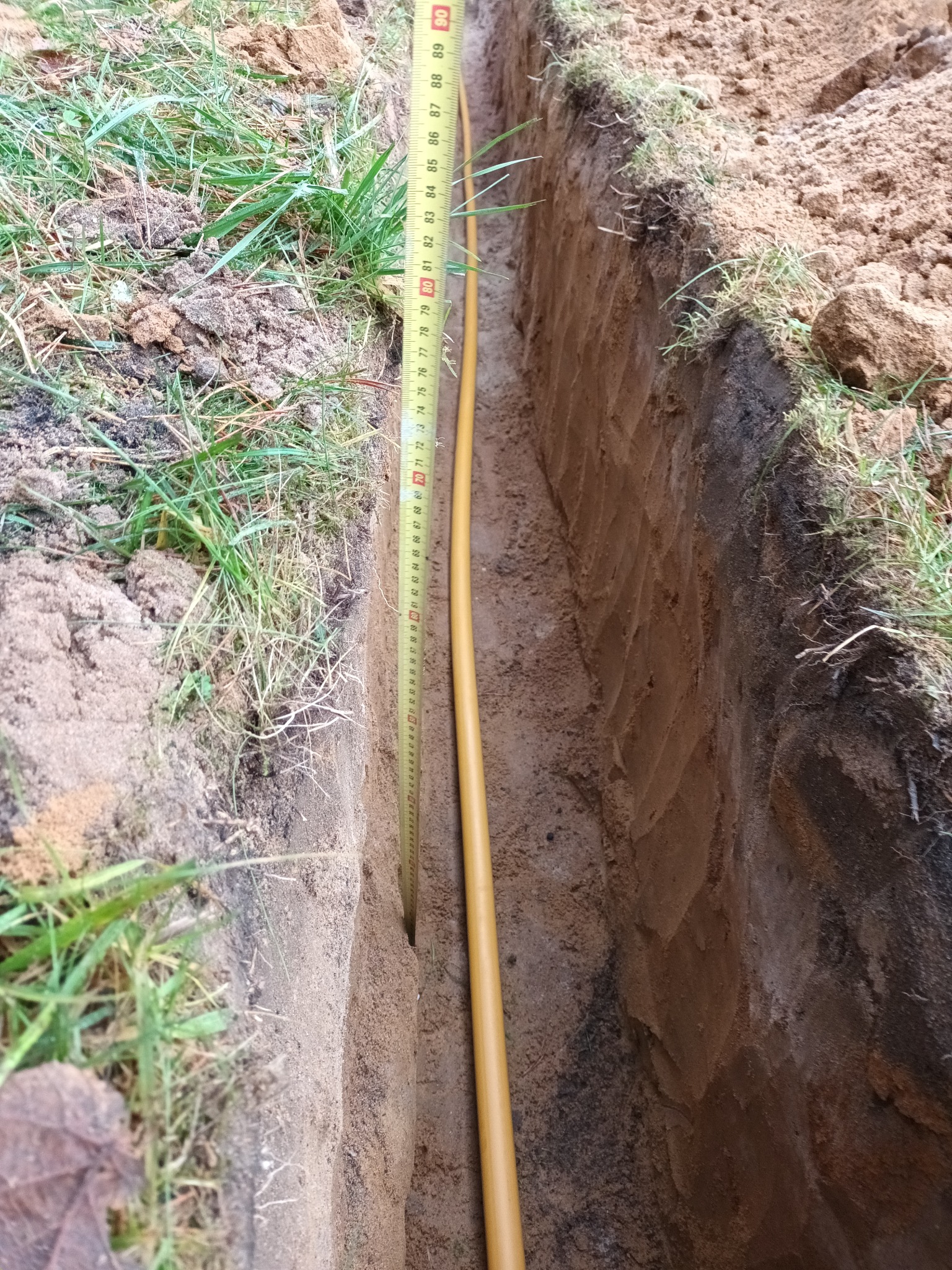 Wykopany wąski rów w ziemi z żółtą rurą na dnie, obok pionowo ustawiona miarka zwijana pokazująca głębokość wykopu.