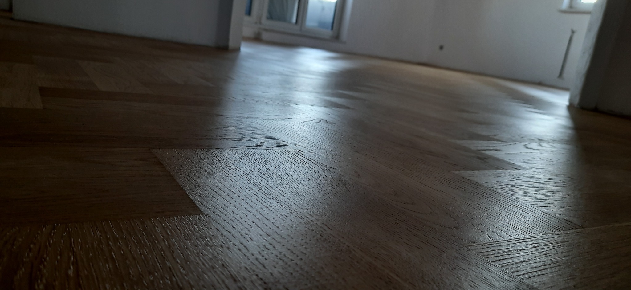 Świeżo ułożony, lakierowany parkiet w jodełkę, z widocznymi promieniami światła odbijającymi się od powierzchni.