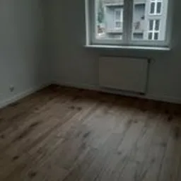 Puste, jasne pomieszczenie z drewnianą podłogą po remoncie, widoczne okno z białymi ramami i fragment budynku za nim.