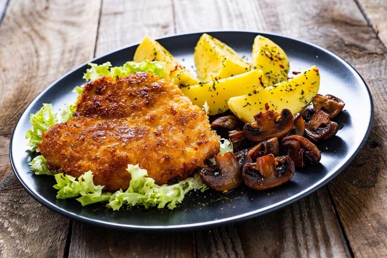 Kotlet schabowy w panierce na sałacie, podany z ziemniakami posypanymi ziołami i smażonymi pieczarkami na czarnym talerzu, rustykalne drewniane tło.