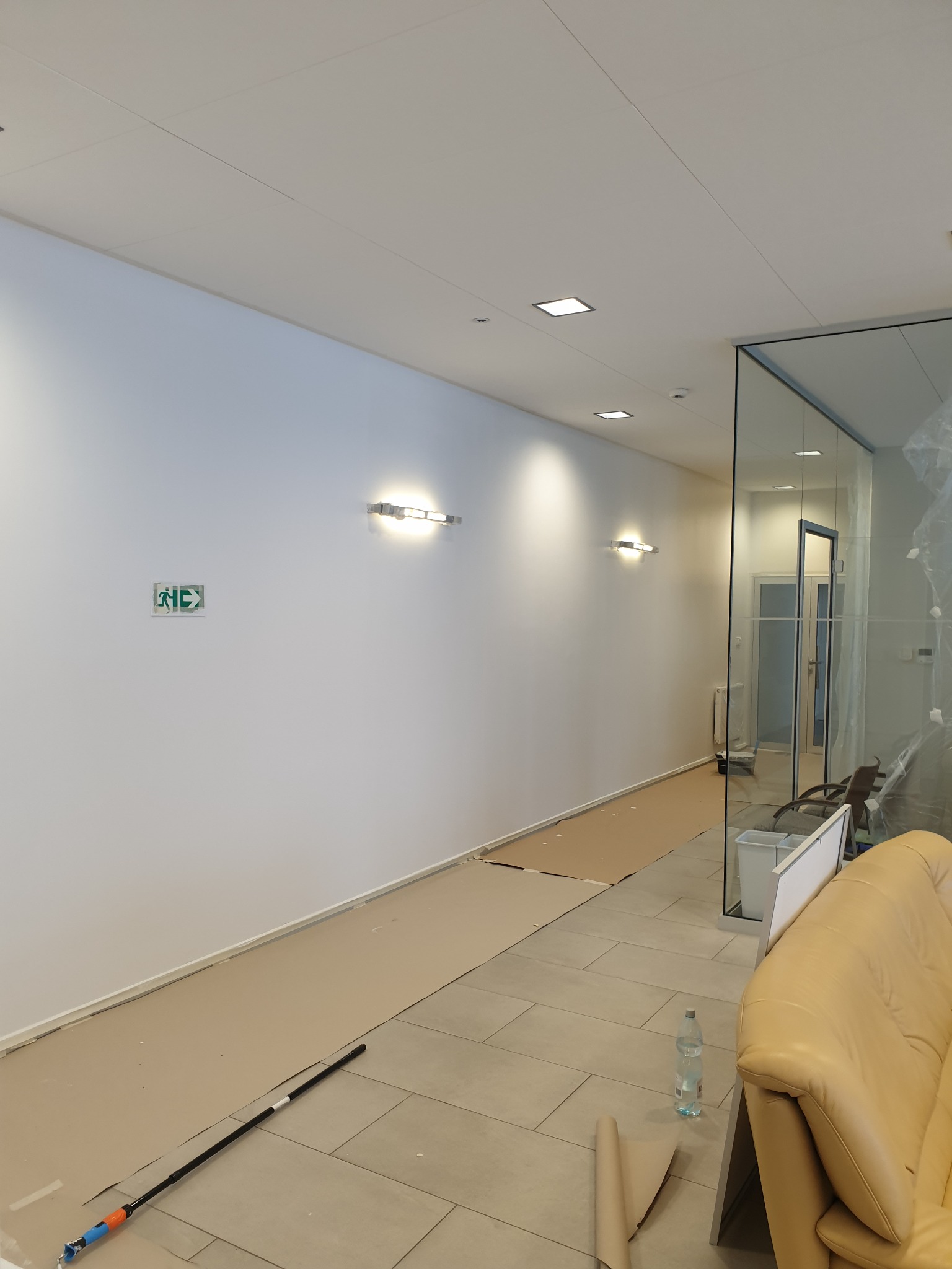 Korytarz biurowy w trakcie malowania, zabezpieczona podłoga kartonem, widoczny wałek do malowania, butelka wody i fragment sofy.