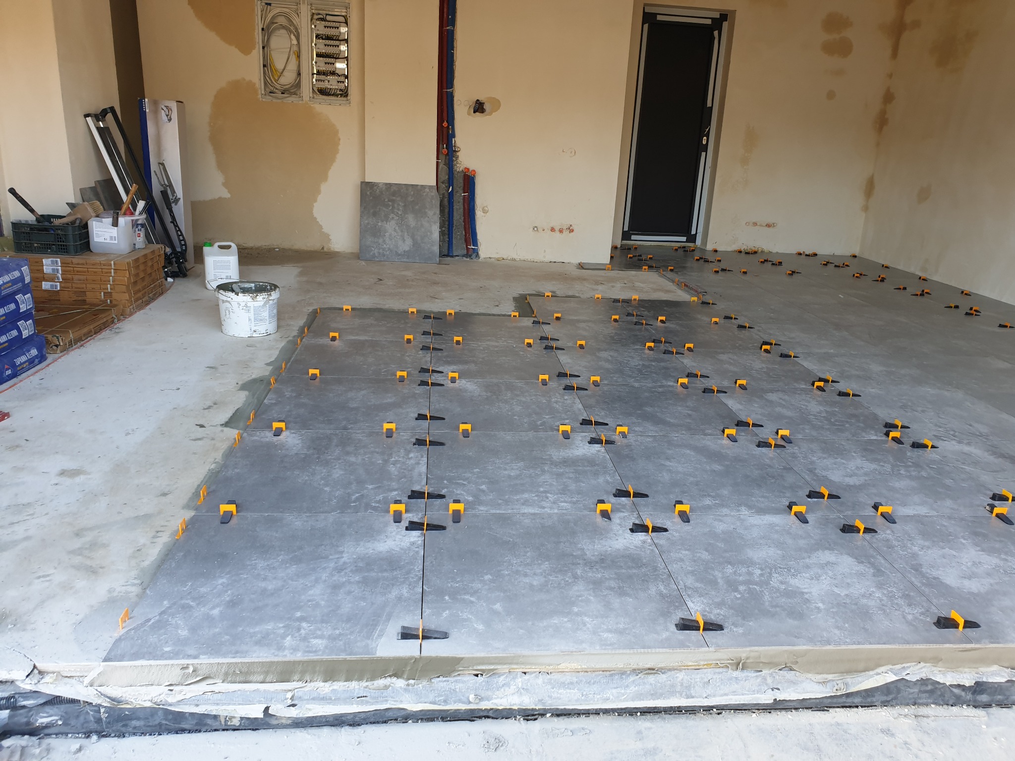 Układanie gresu na podłodze z użyciem systemu poziomowania płytek, widoczne narzędzia i materiały budowlane w tle.