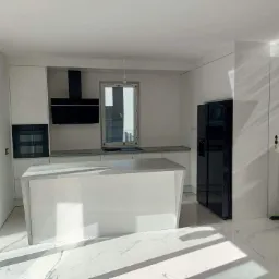 Minimalistyczna, jasna kuchnia z wyspą i czarnymi akcentami, częściowo wykończona, z widocznym sprzętem AGD i marmurową podłogą.