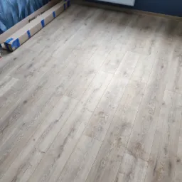 Ułożone panele podłogowe w jasnym odcieniu drewna, widoczne opakowanie po panelach przy ścianie w pokoju z niebieskimi ścianami i białym grzejnikiem.