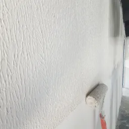 Nakładanie białej, dekoracyjnej struktury na ścianę za pomocą wałka malarskiego z pomarańczową rączką; widoczna faktura ściany przypominająca korę drzewa.