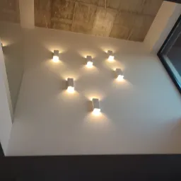 Biała ściana z siedmioma kinkietami w kształcie prostopadłościanów, betonowy sufit i fragment okna z widocznym niebieskim niebem.