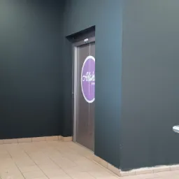 Świeżo pomalowana ciemnoszara ściana korytarza z widocznymi śladami malowania przy listwach przypodłogowych, obok wejście do windy z naklejką w kształcie fioletowego koła.