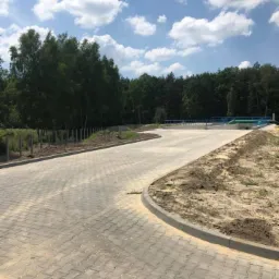 Szeroki, wybrukowany plac z kostki w odcieniach szarości, otoczony krawężnikiem i przygotowanym pod nasadzenia terenem, w tle widoczne drzewa i błękitne niebo z białymi obłokami.