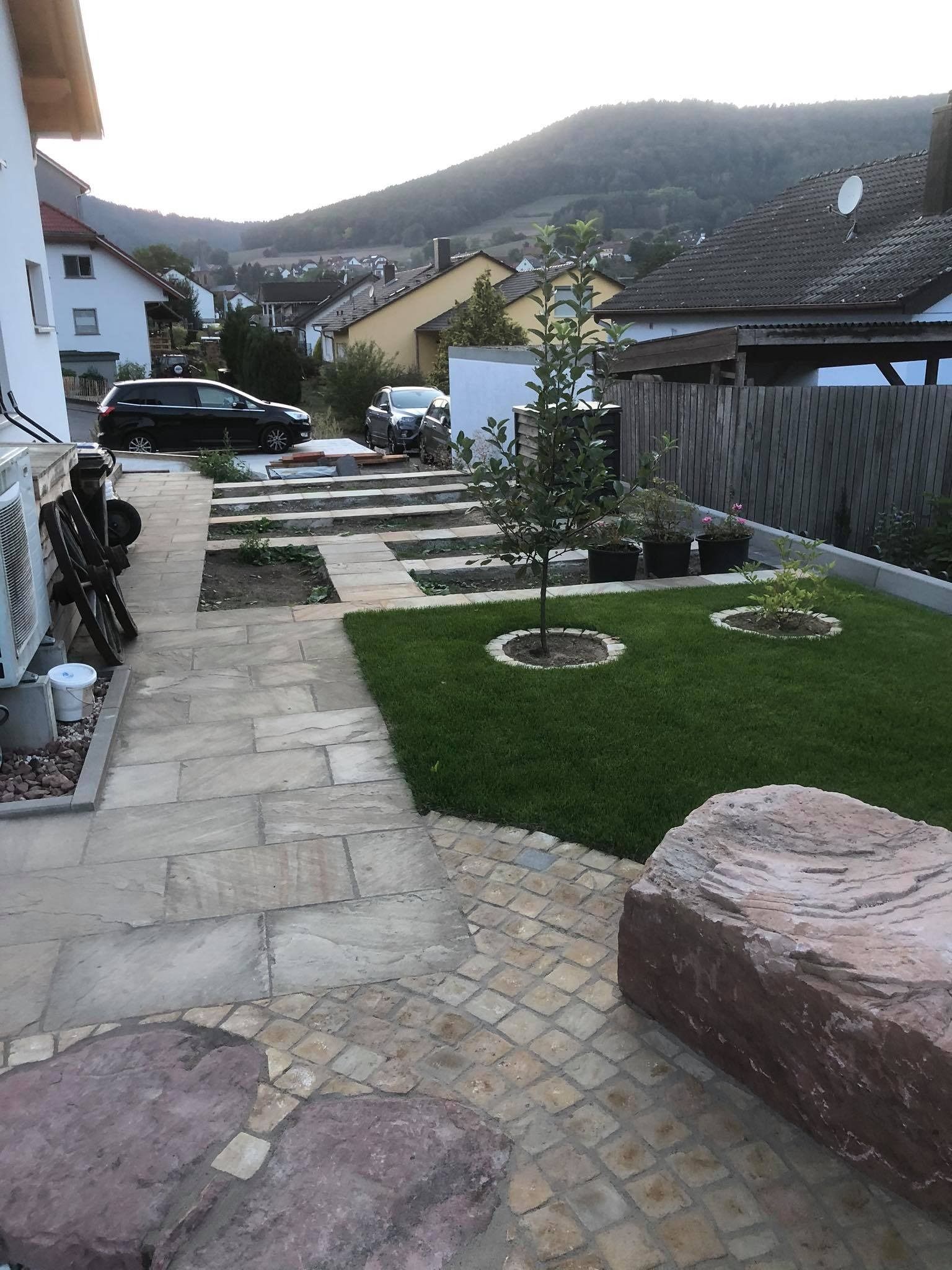 Ogród z kamienną ścieżką, trawnikiem z młodymi drzewkami i widokiem na zabudowania w tle