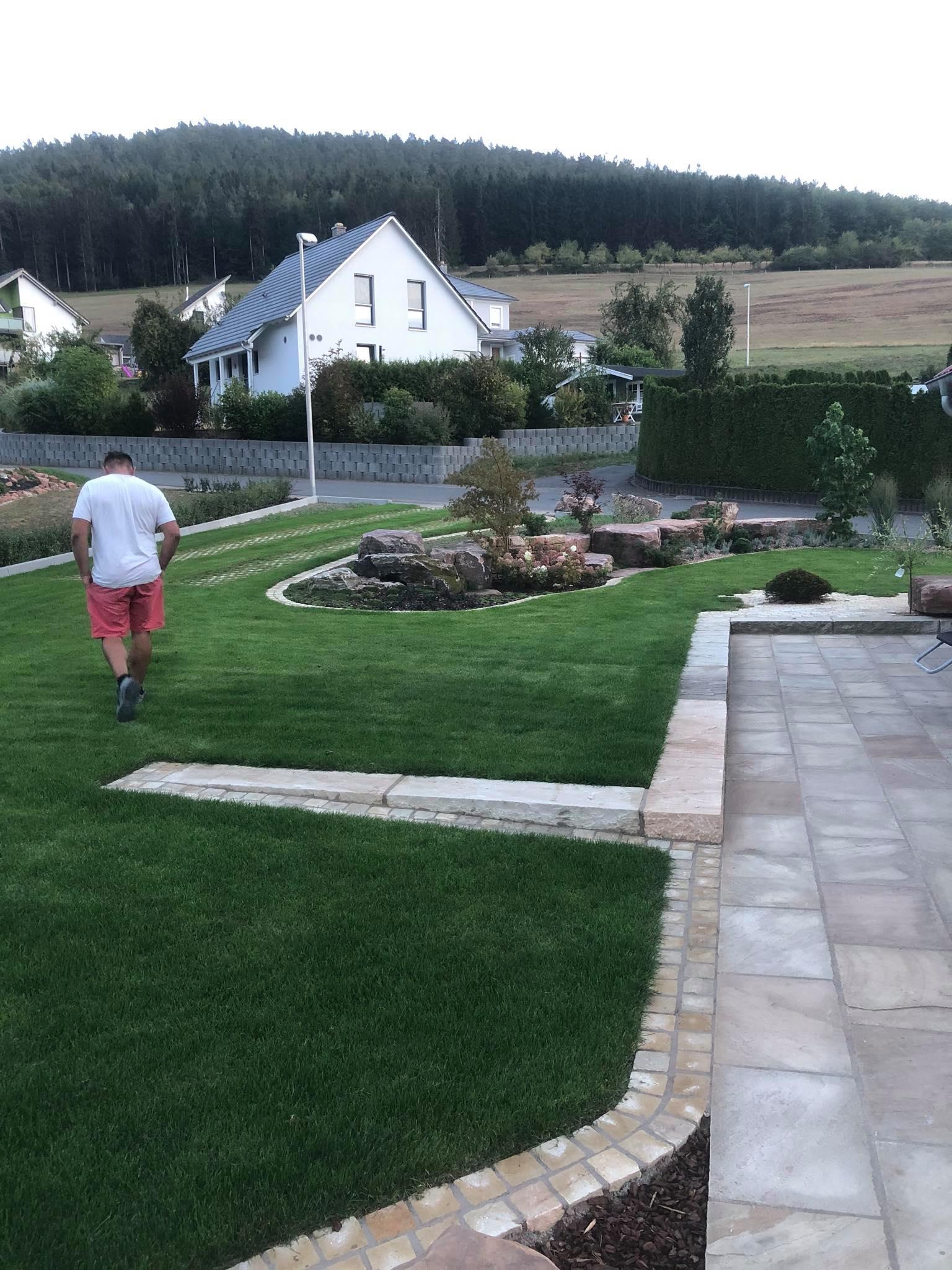 Ogród z nową nawierzchnią z kostki brukowej i trawnikiem, widok zza pleców mężczyzny w różowych spodenkach.