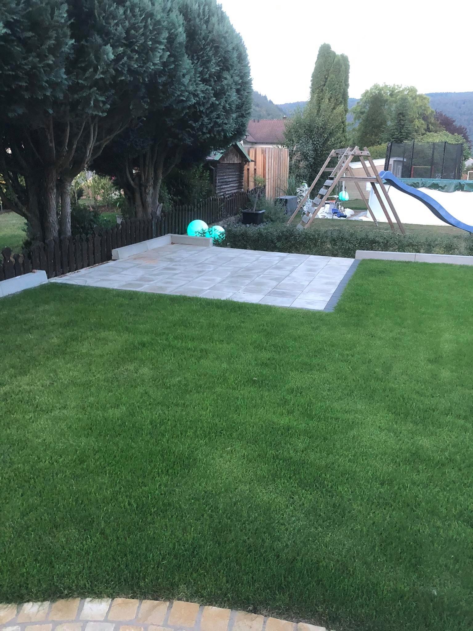 Wykończony taras z jasnej kostki brukowej na trawniku, w tle drzewa i plac zabaw z huśtawką i zjeżdżalnią.