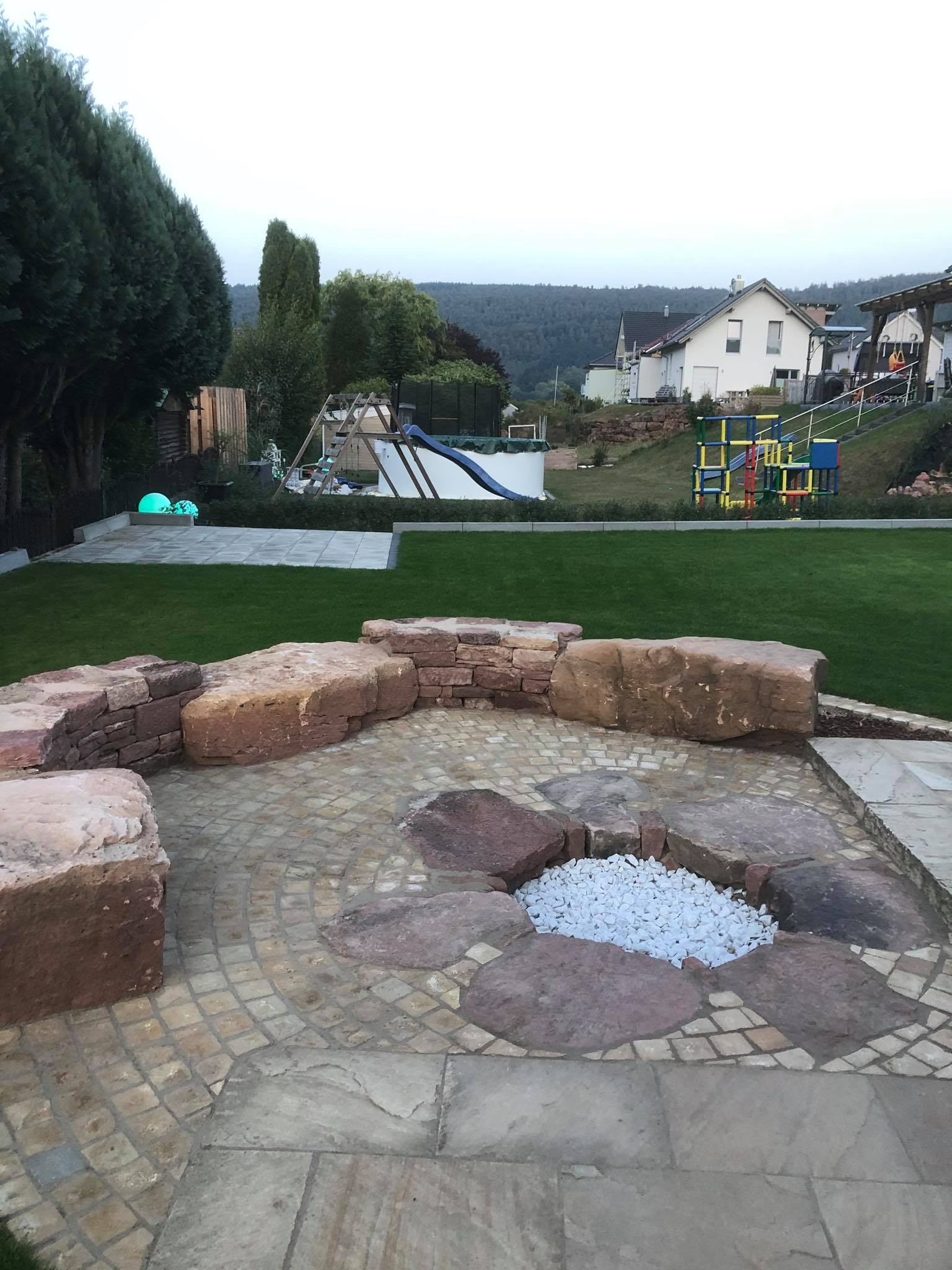 Ogród z paleniskiem otoczonym kamiennymi blokami i ścieżką z drobnej kostki brukowej, w tle trawnik i elementy placu zabaw.