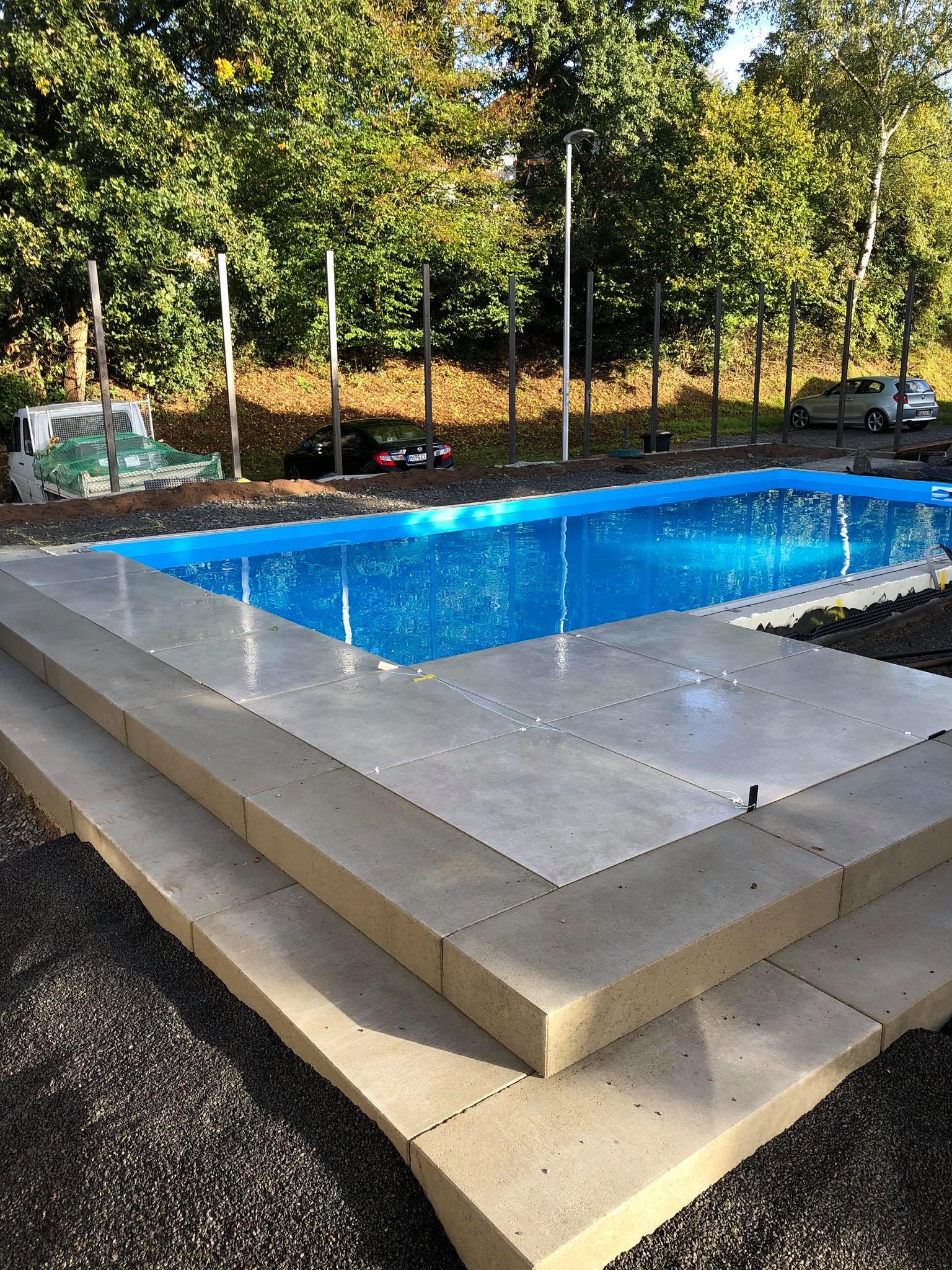 Niebieski basen z wykończeniem z jasnych płyt, widoczne schody i czarna podsypka, w tle zieleń drzew i zaparkowane samochody.