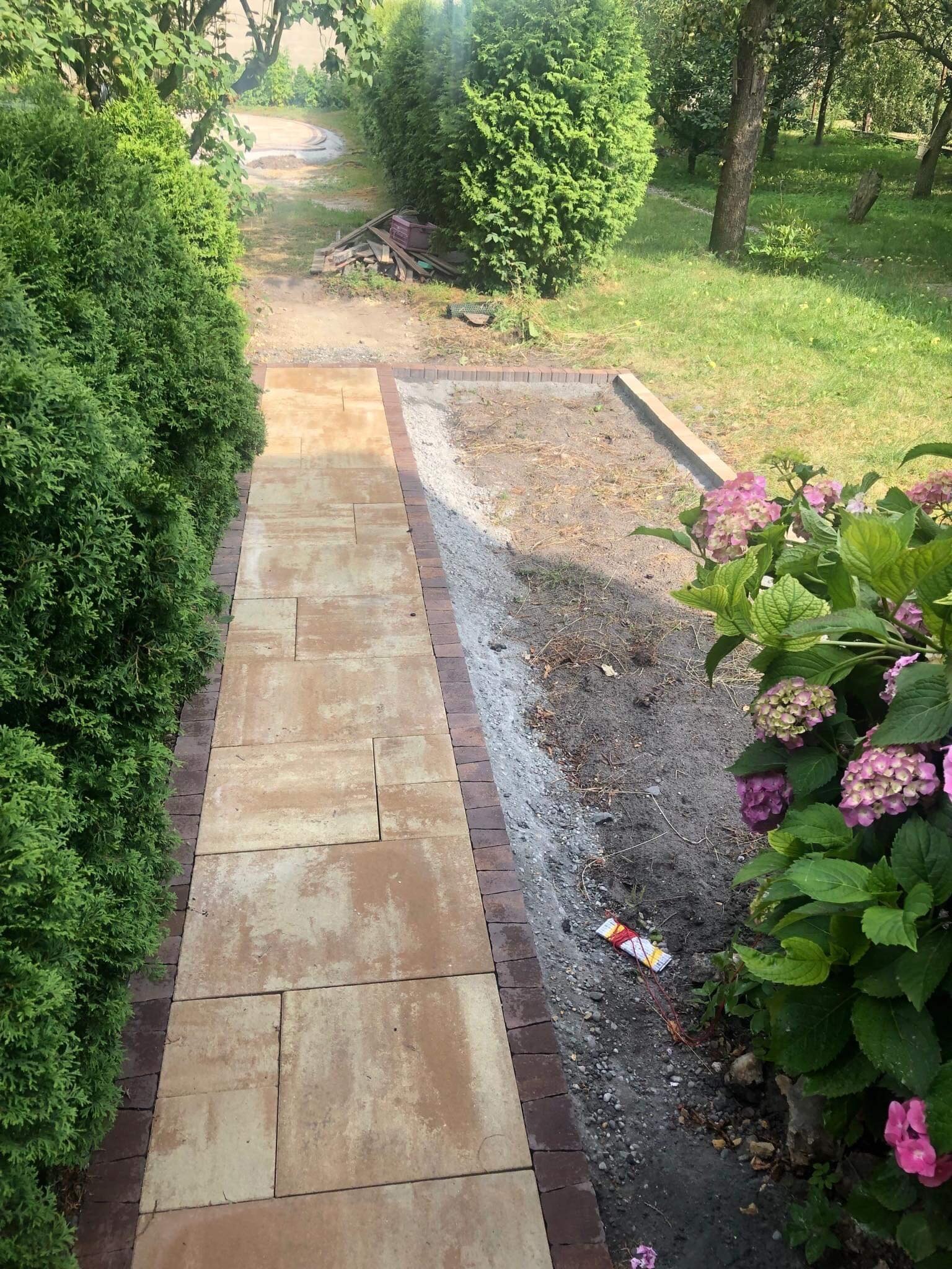 Ułożona ścieżka z beżowej kostki brukowej obramowana czerwoną kostką, widok z góry, obok rosną tuje i hortensje, w tle trawnik i drzewa.
