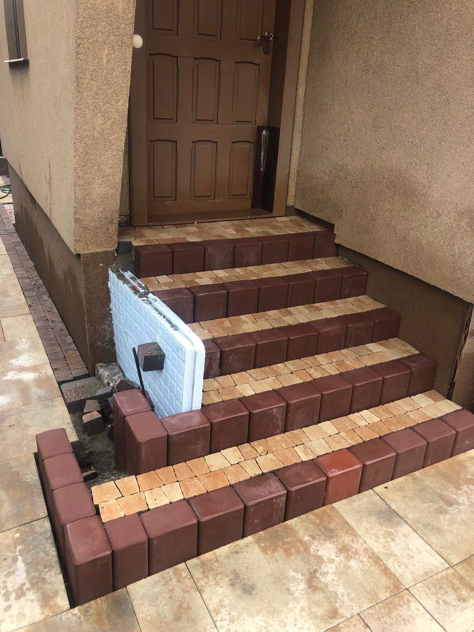 Schody wejściowe do domu w trakcie budowy z kostki brukowej w odcieniach brązu i beżu, widoczna izolacja termiczna ściany fundamentowej