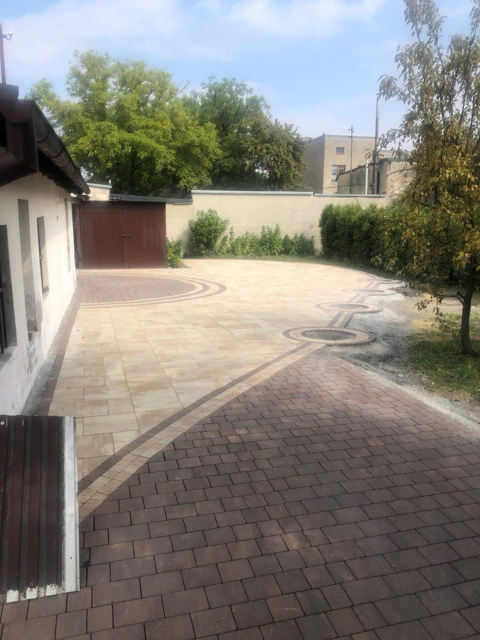 Ułożona kostka brukowa w dwóch odcieniach brązu i beżu tworząca wzór z okręgami na podjeździe przed budynkiem z drewnianą bramą garażową.