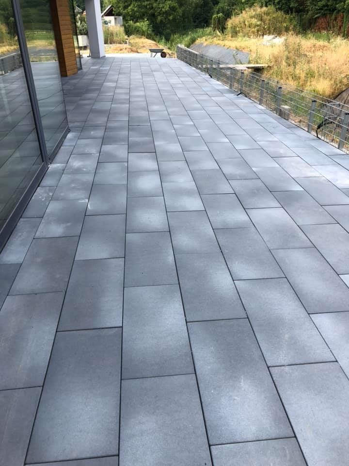 Ułożona szara kostka brukowa na tarasie przylegającym do budynku z dużymi oknami, widoczne tło z zielenią i w oddali taczka.