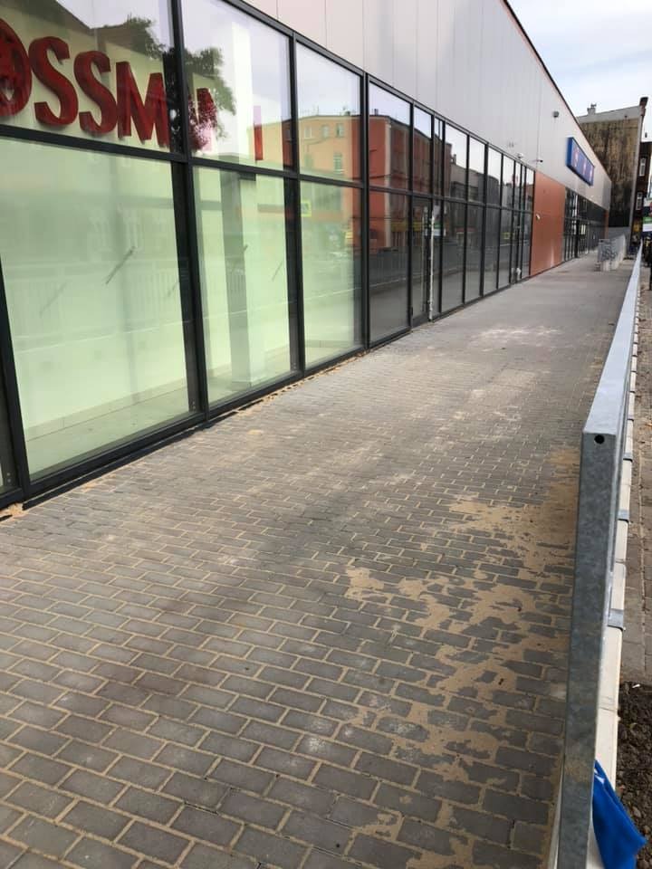 Nowo ułożona, szara kostka brukowa tworząca chodnik i podjazd dla wózków inwalidzkich przed budynkiem sklepowym z dużymi oknami.