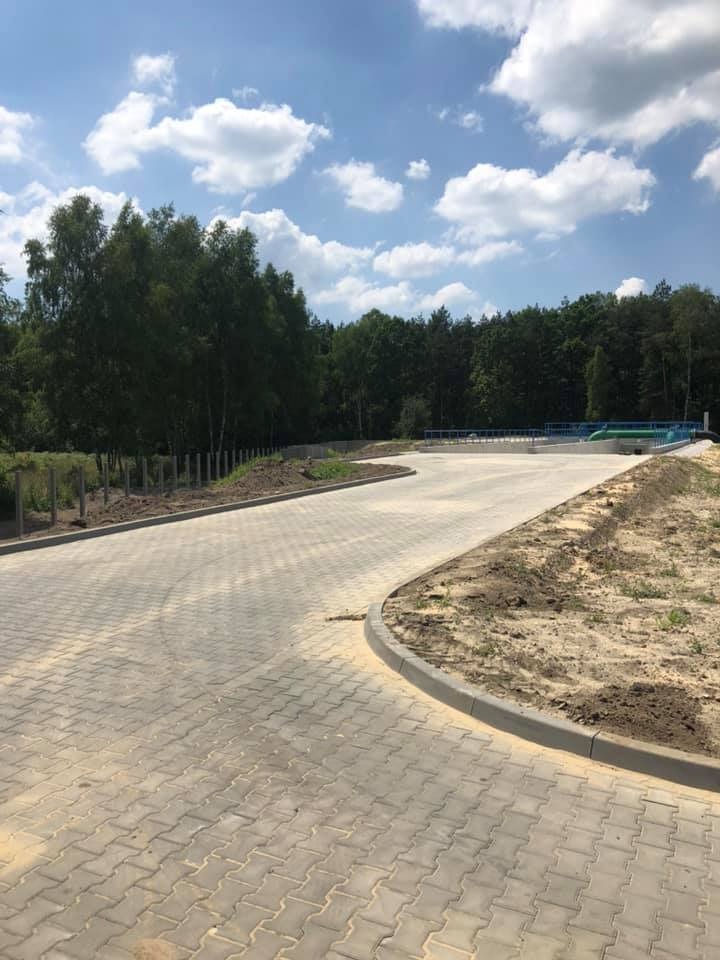 Szeroki, wybrukowany plac z kostki w odcieniach szarości, otoczony krawężnikiem i przygotowanym pod nasadzenia terenem, w tle widoczne drzewa i błękitne niebo z białymi obłokami.