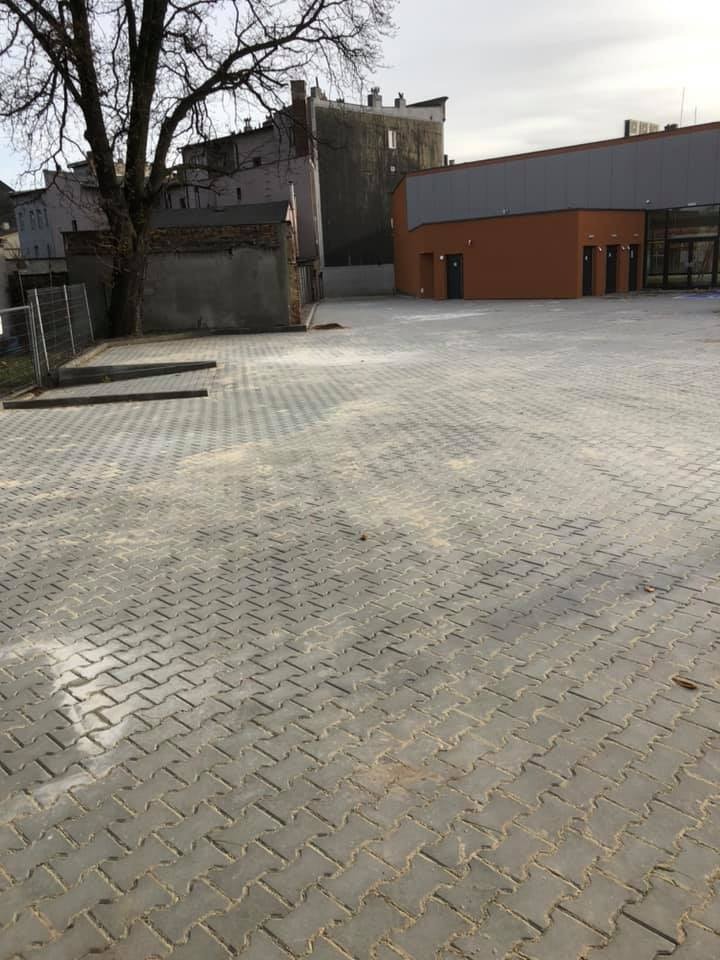 Ułożona szara kostka brukowa na dużym placu, widoczne budynki w tle i nagie drzewo po lewej stronie, lekkie zabrudzenia na kostce.