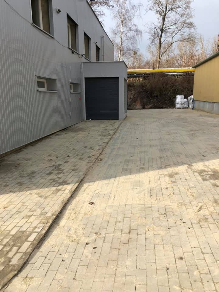 Wyłożony kostką brukową podjazd do garażu blaszanego, widoczne ślady piasku na powierzchni, budynek przemysłowy w tle.