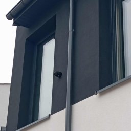 Woj-Bol Technology Wojciech Ból - Kamera monitoringu zamontowana na elewacji budynku w pobliżu okna. Widoczny fragment dachu, rynna i dwukolorowa elewacja. Ujęcie z dołu.