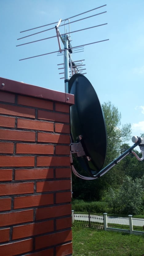 Antena satelitarna i antena naziemna zamontowane na kominie z czerwonej cegły, widok na błękitne niebo i zieleń ogrodu.
