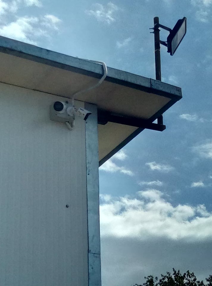 Biała kamera monitoringu zewnętrznego przymocowana do rogu budynku z blachy, z widocznymi przewodami i lampą oświetleniową na słupie w tle, na tle błękitnego nieba z chmurami.