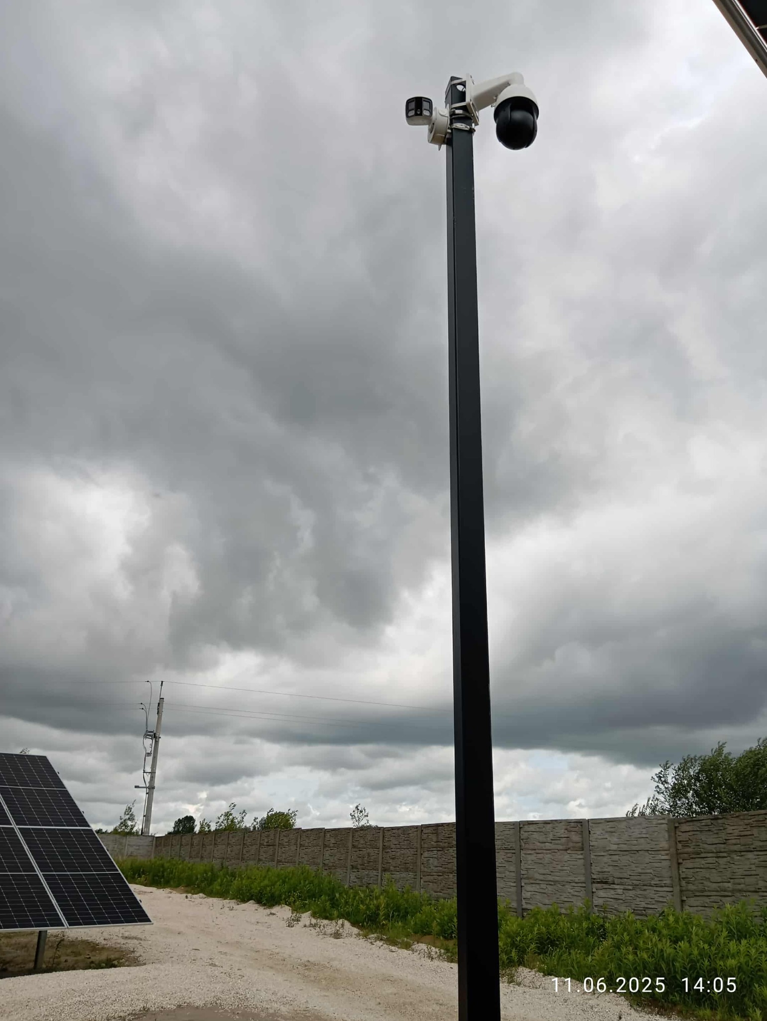 Zewnętrzny system monitoringu na słupie z kamerą obrotową i dodatkową kamerą, na tle pochmurnego nieba i paneli słonecznych. Widoczny betonowy płot.