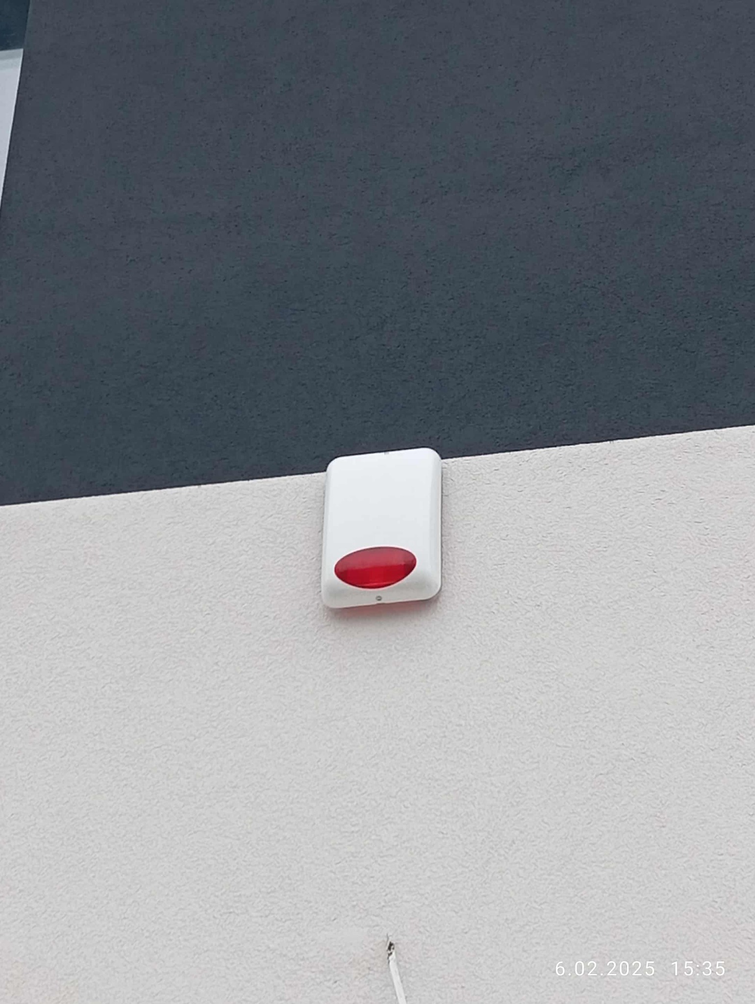 Biała obudowa sygnalizatora alarmowego z czerwoną diodą, zamontowana na dwukolorowej elewacji budynku. Widoczne okablowanie.