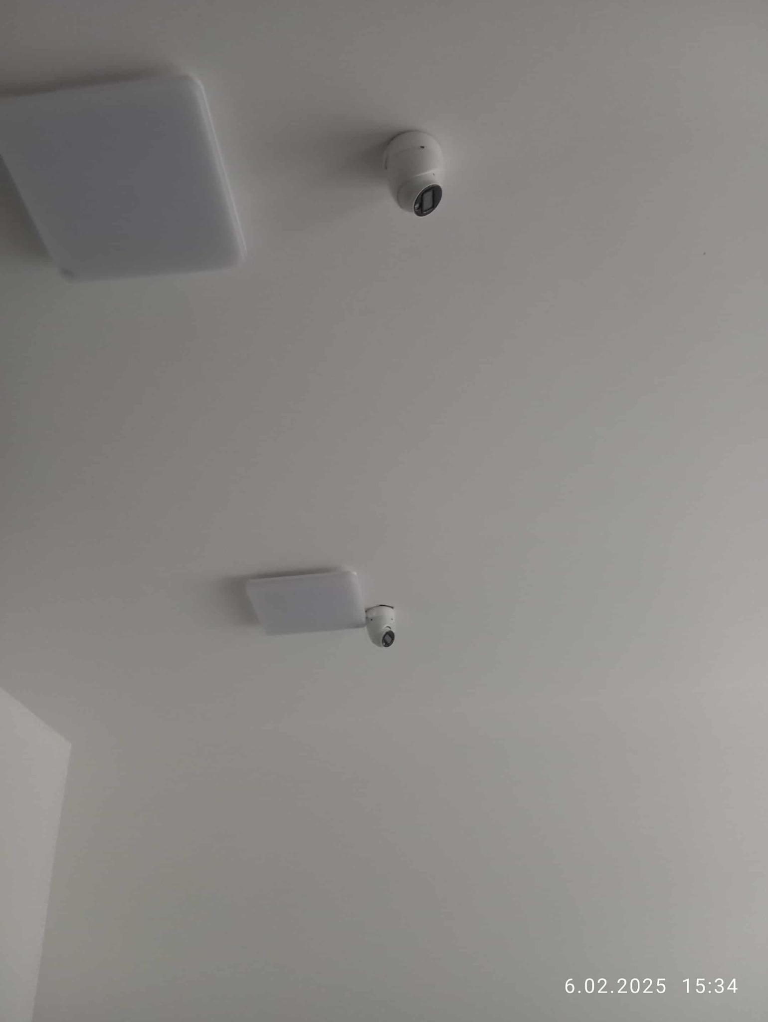 Biały sufit z dwoma lampami LED i dwiema kamerami monitoringu, skierowanymi w dół. Minimalistyczny design, neutralne oświetlenie.
