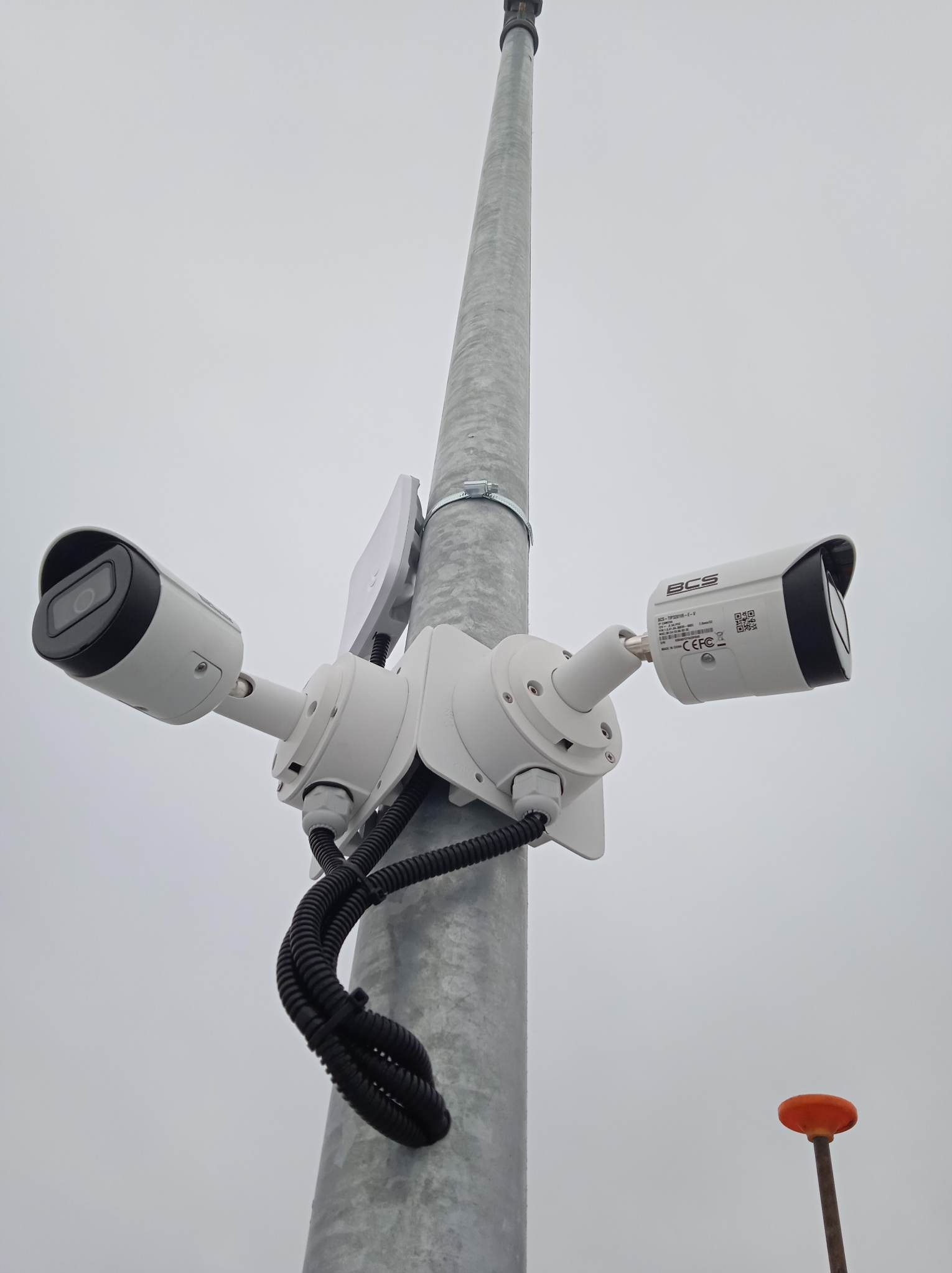 Dwie kamery monitoringu BCS zamontowane na metalowym słupie, skierowane w przeciwne strony. Widoczne przewody i uchwyty montażowe. Szare niebo w tle.