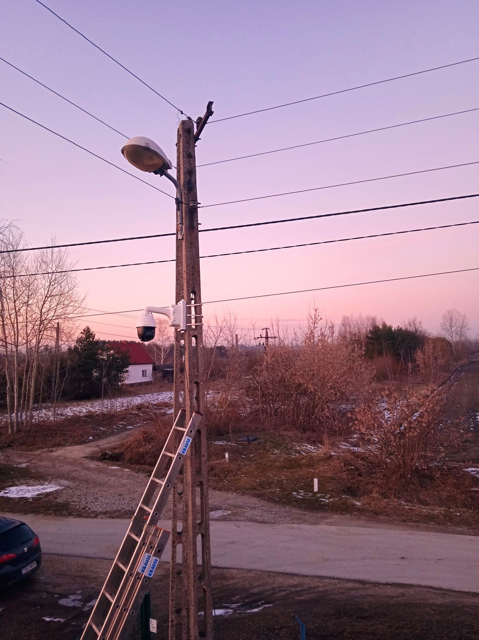 Kamera monitoringu PTZ zamontowana na słupie energetycznym w Pacanowie, widoczna drabina Krause. Tło: zachodzące słońce i zabudowa jednorodzinna.