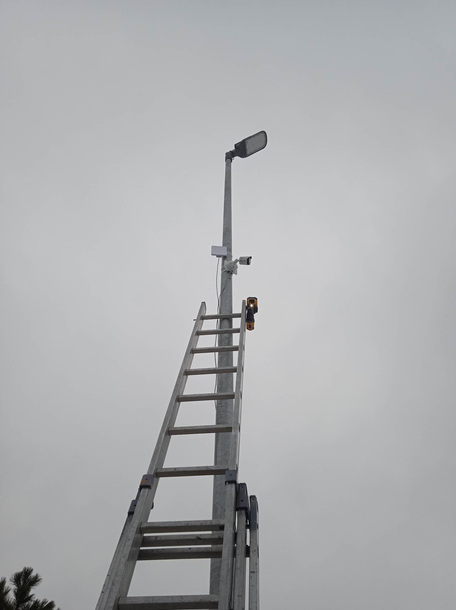 Słup oświetleniowy z zamontowaną kamerą monitoringu, drabina przystawiona do słupa. Widok z dołu na szarym tle nieba. Sprzęt instalacyjny na szczeblach.