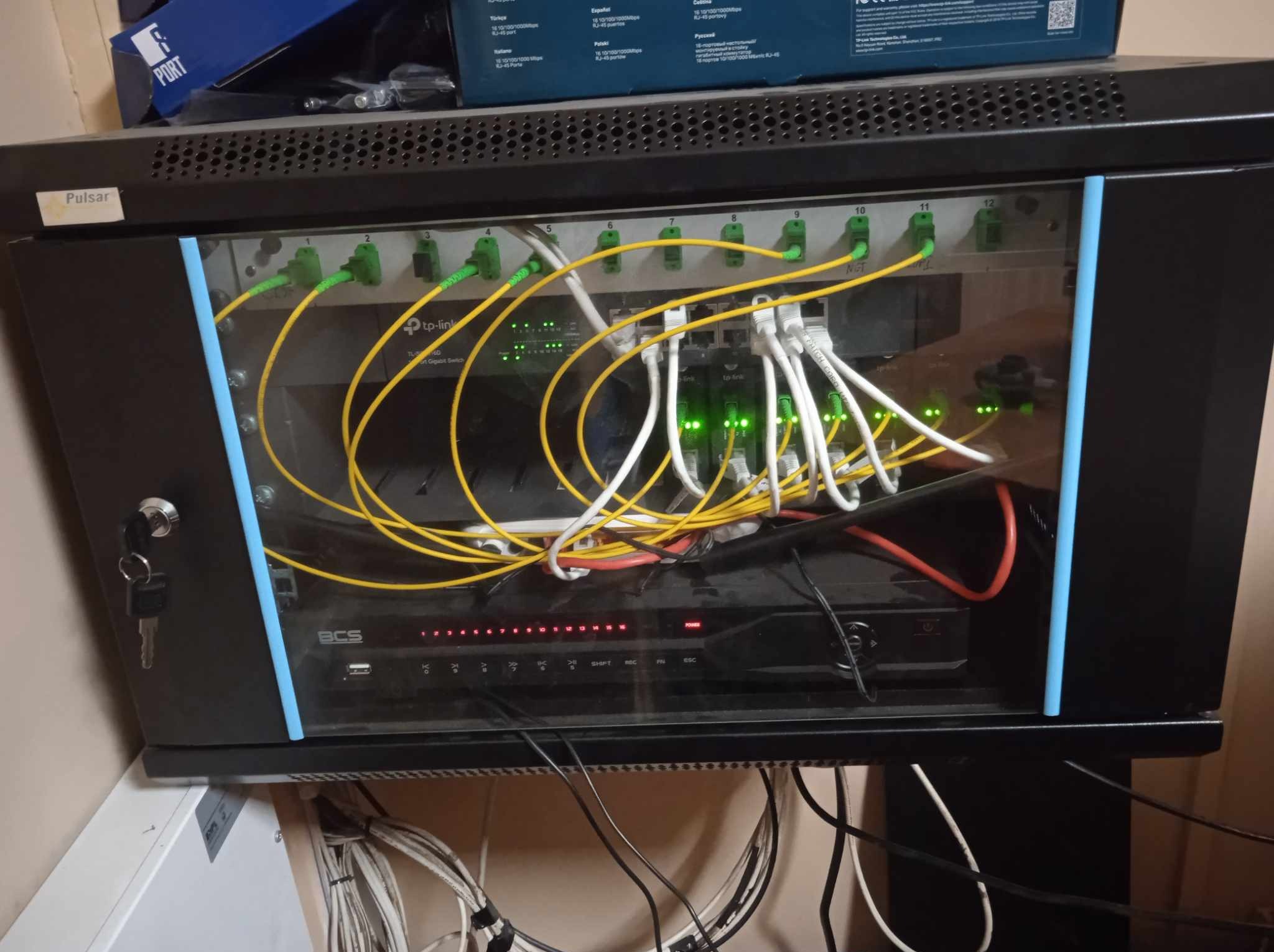 Szafa rack z urządzeniami sieciowymi: switch TP-Link, rejestrator BCS. Widoczne kable światłowodowe i ethernetowe. Klucze w zamku szafy.