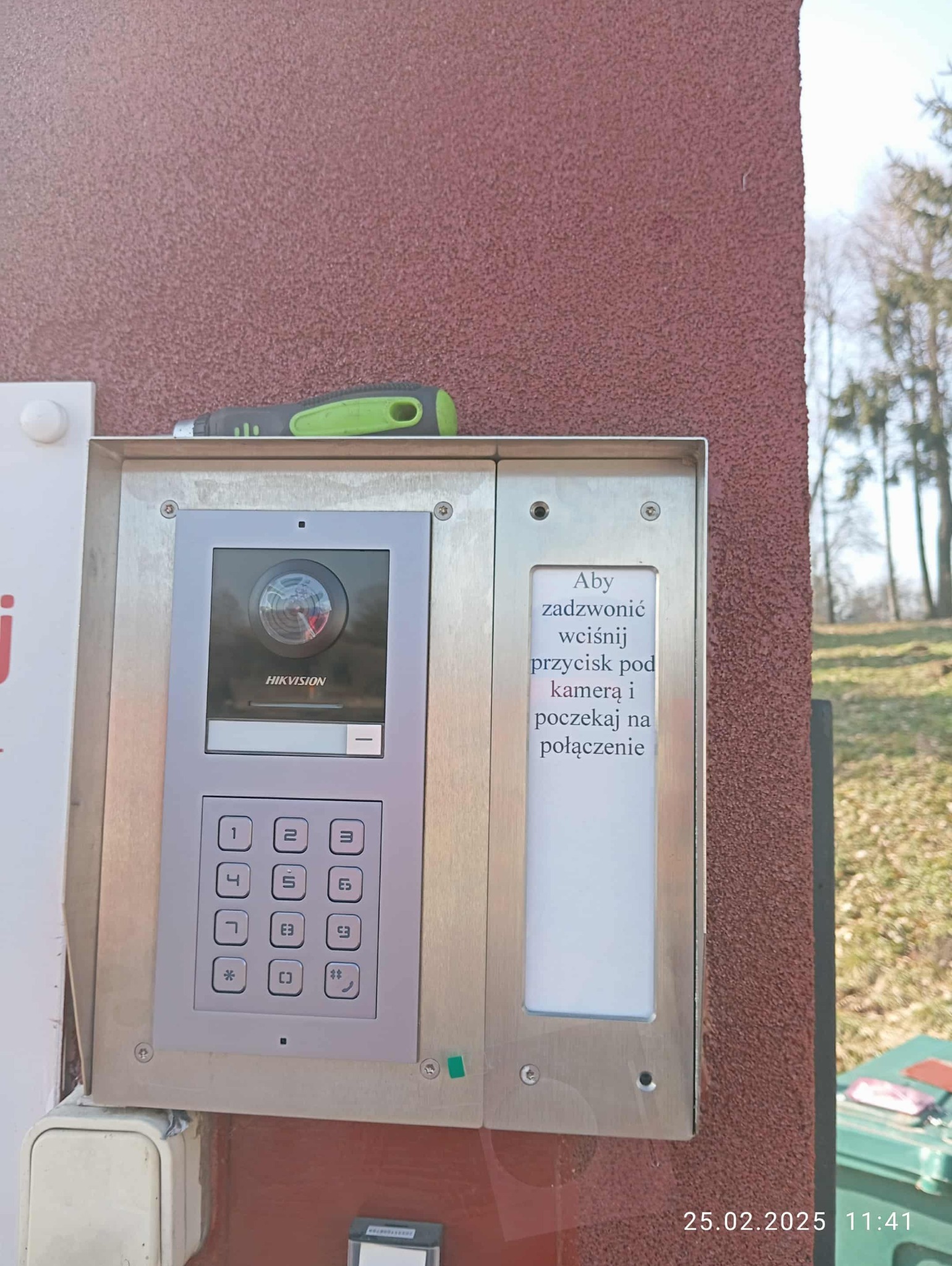 Zamontowany wideodomofon Hikvision z kamerą i klawiaturą numeryczną na elewacji budynku. Nad urządzeniem leży śrubokręt. Widoczna instrukcja obsługi.