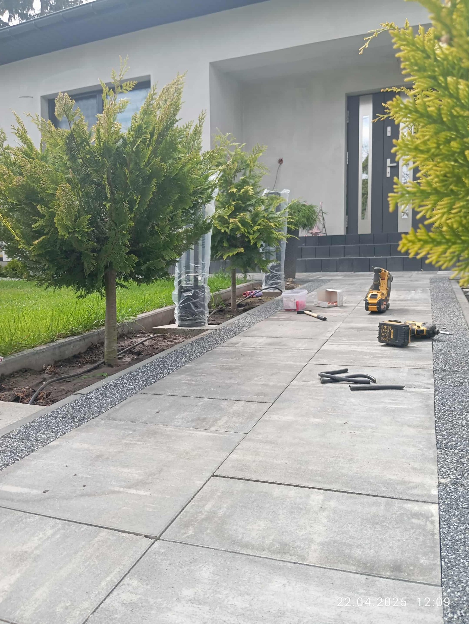 Ułożona kostka brukowa przed domem z posadzonymi drzewkami. Widoczne narzędzia i kable, sugerujące prace instalacyjne w toku. Jasne, dzienne światło.