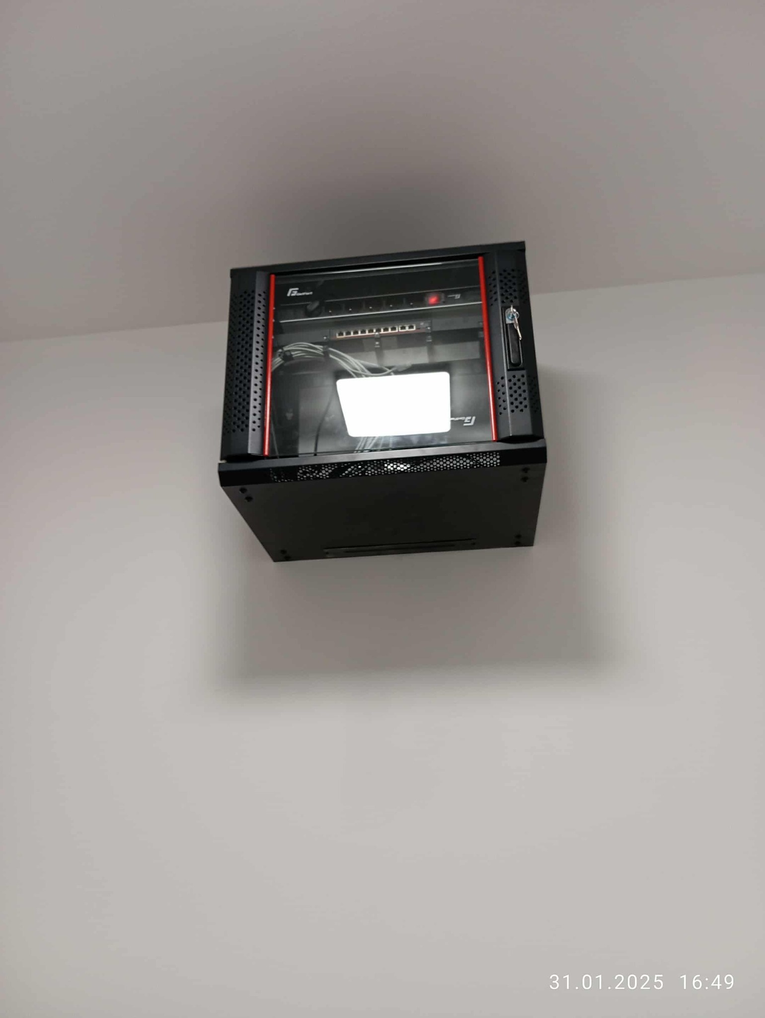 Czarna szafa rack z czerwonymi akcentami, zamontowana na białej ścianie. Widoczne kable i urządzenia wewnątrz. Kąt ujęcia od dołu.