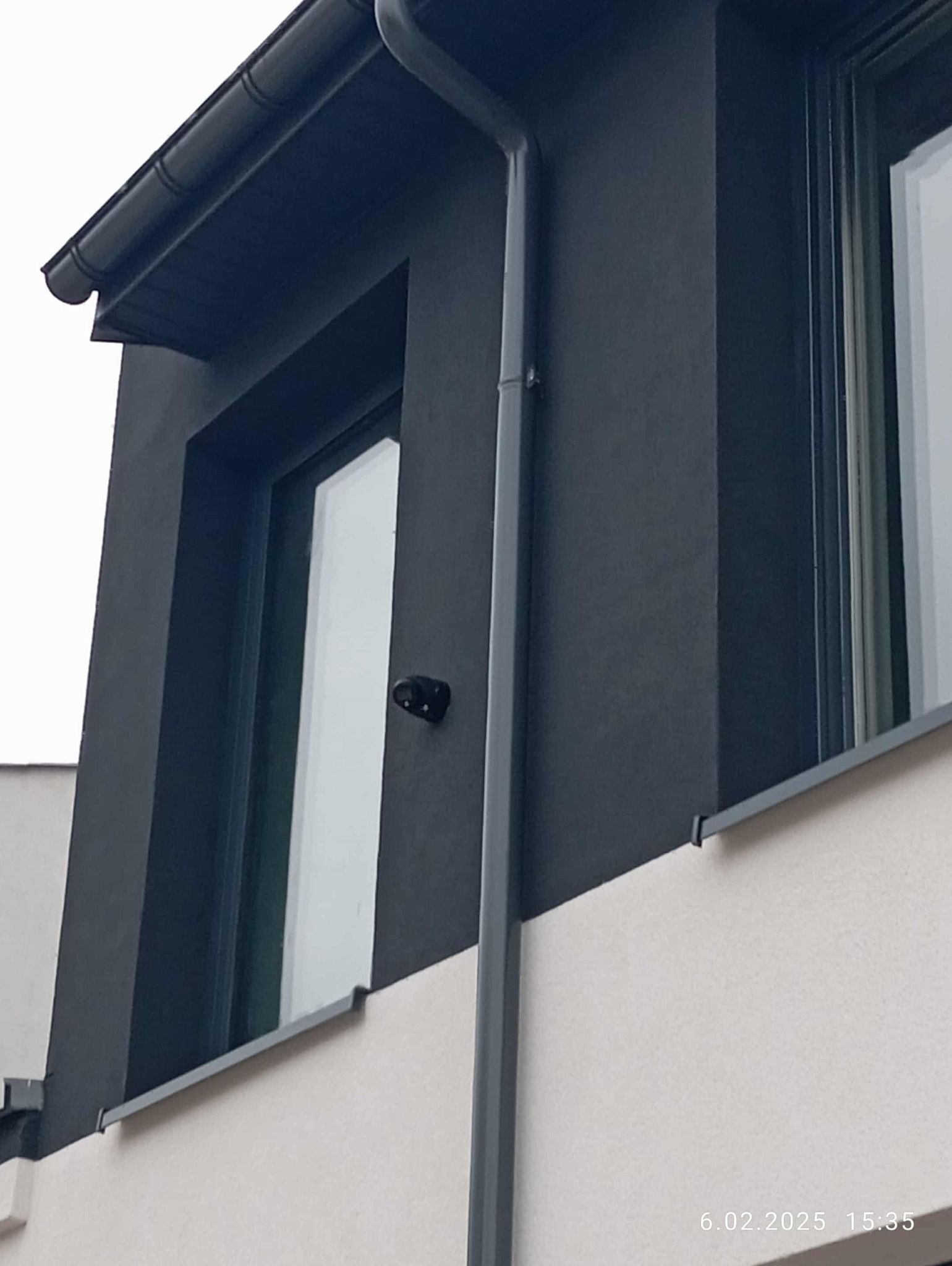 Kamera monitoringu zamontowana na elewacji budynku w pobliżu okna. Widoczny fragment dachu, rynna i dwukolorowa elewacja. Ujęcie z dołu.