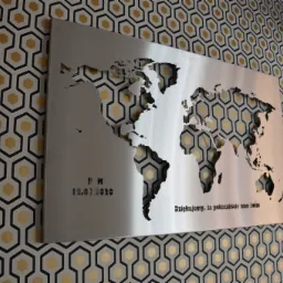 Metalowa mapa świata wycięta laserowo, zamontowana na ścianie z geometryczną tapetą. Widoczne napisy i data wycięte w metalu.