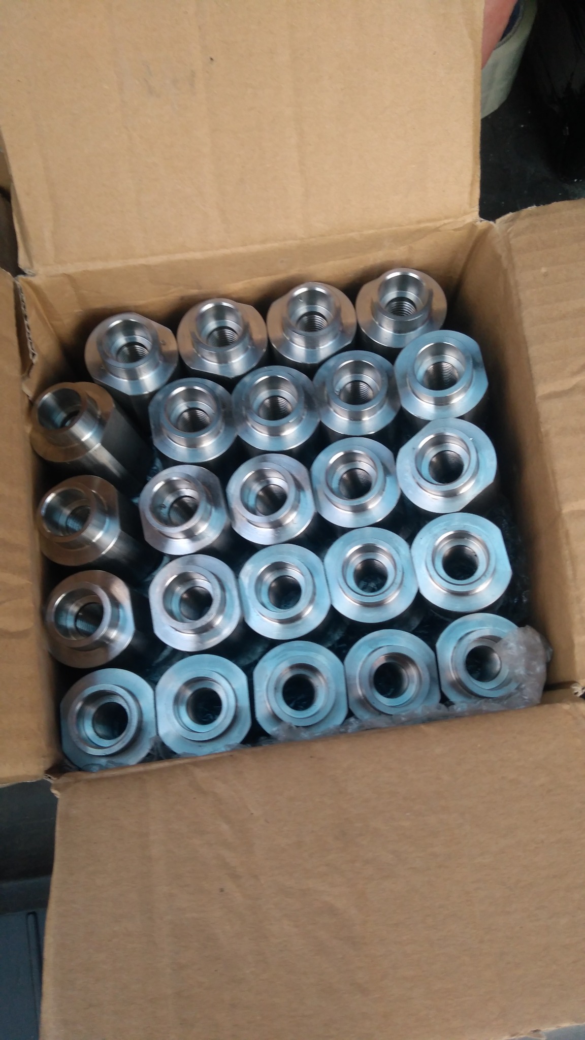 Partia metalowych tulei z gwintem wewnętrznym, ułożonych rzędami w kartonowym pudełku, widoczne ślady obróbki skrawaniem.