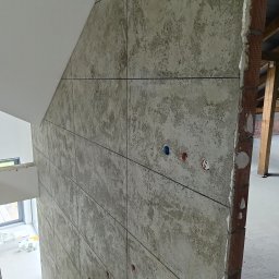 LukUp - Ściana wykończona płytami imitującymi beton, z widocznymi puszkami elektrycznymi. Surowy, industrialny styl wykończenia wnętrza w trakcie realizacji. Widoczne schody i ceglany fragment ściany.