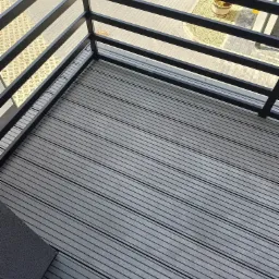 Szare deski kompozytowe na balkonie, widok z góry. Czarne, metalowe barierki i fragment widoku na zewnątrz z rośliną w donicy. Nowoczesna aranżacja balkonu.