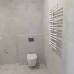 Nowoczesna łazienka w jasnych odcieniach szarości z wiszącą toaletą i białym grzejnikiem drabinkowym, wykończona płytkami imitującymi kamień.