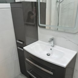 LukUp - Nowoczesna łazienka z szarą szafką pod umywalkę, lustrem z podświetleniem LED i wysoką szafką. Białe płytki imitujące marmur na ścianach i szara podłoga.