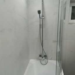 LukUp - Biała, wykończona łazienka z wanną, szklanym parawanem i chromowaną armaturą. Ściany wyłożone płytkami imitującymi marmur. Widok na kabinę prysznicową.